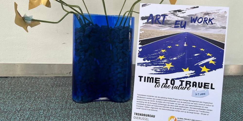 Een blauwe vaas met stenen en gouden bloemen staat naast een flyer met de titel "ART EU WORK TIME TO TRAVEL to the future". De flyer informeert over een evenement van studenten Global Project and Change Management aan Hogeschool Windesheim, over de toekomst van werk, kunst, cultuur en de EU.
